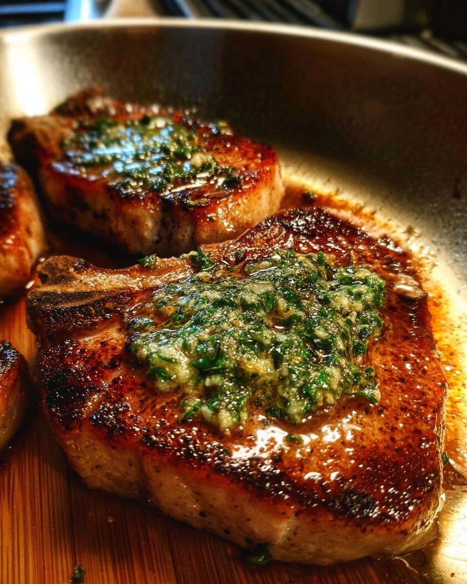 Close-up of juicy From Scratch Pork Chops With Herb Butter sizzling in a pan, topped with vibrant green herb butter.